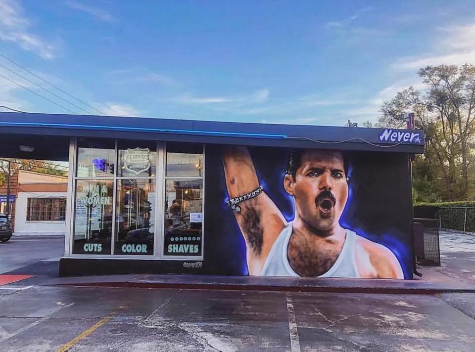 There is a shop with a sloped blue roof and on the front wall next to a shop window, there is a painting mural of Freddie Mercury with his one arm up, a black armband on it, and his mouth shaped like an o and his black hair slicked back. This is his iconic pose from his Wembley performance.