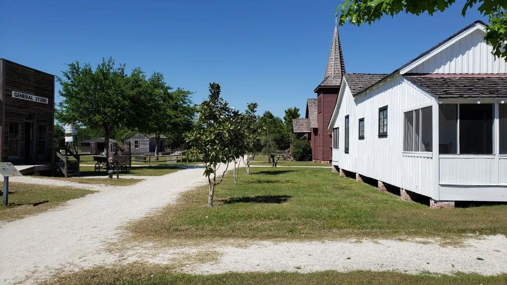 Deserted street with a white house on the corner and behind there is a grey building which has a pointy roof, looks like a church. There are small trees on the side of dirt track road.