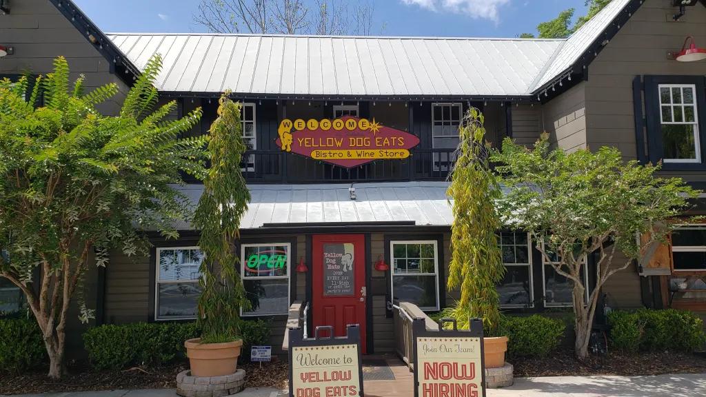 Brown building with a red and yellow sign saying "Yellow Dog Eats". There are two signs outside saying "eat no2 at Yellow Dog Eats" and "now hiring" with two trees on either side.