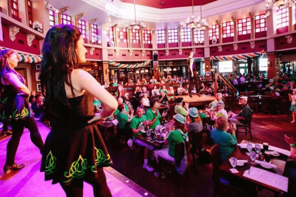 Girls stood on stage in black dresses with green details and black tights on. Below the stage there are loads of tables full of people watching them dance. The pub space is big and open.