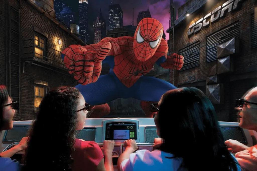 4 people sat on ride vehicle. They are wearing black 4D glasses and it looks like a man stood on the front of the vehicle in spiderman suit. The suit is red with black spider webs on and blue legs.