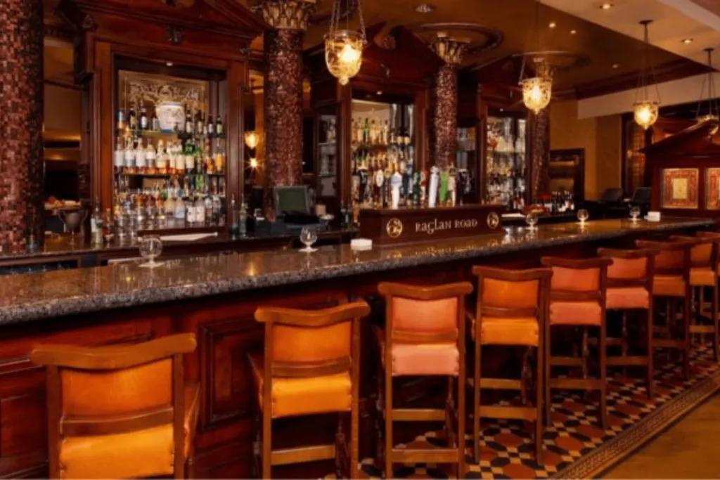 Dark wooden bar with orange and wooden bar stools. There are drinks hanging behind the bar and it is completely empty.