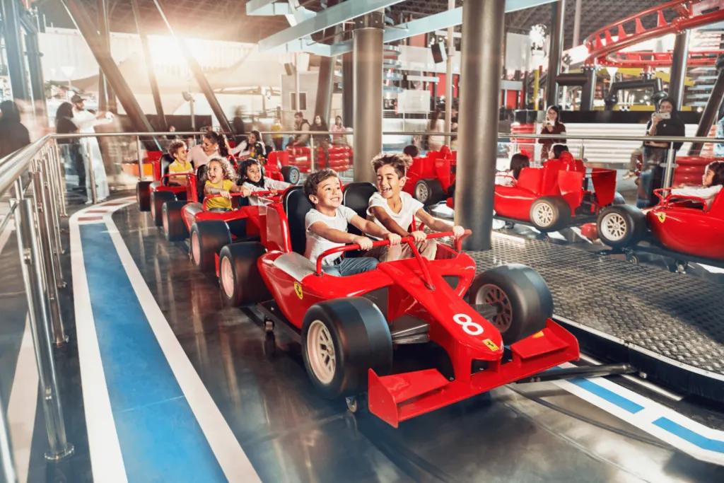 Little ride with small ferrari's lined up on a track. There are small kids sat on this ride holding onto pretend steering wheels.