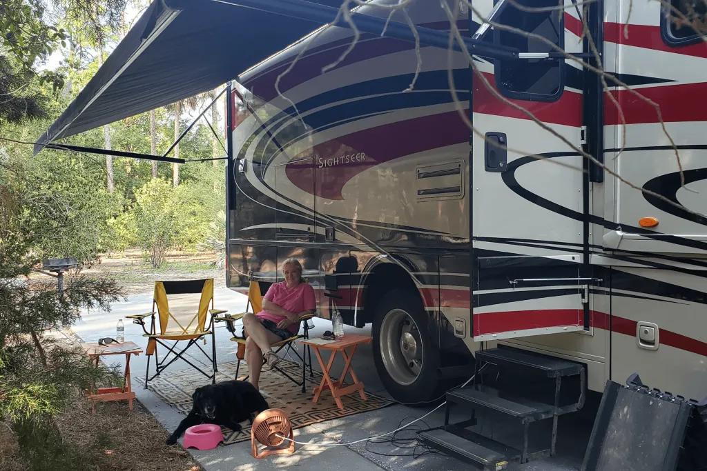 Woman sat on foldable chairs outside a campervan under some shade. The door is open and the black dog is sitting at the woman's feet.