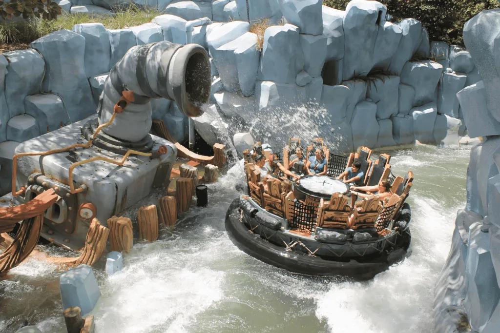 12 people sat in circular raft with wooden seats, going along river rapids surrounded by stone walls and getting splashed from fountains.
