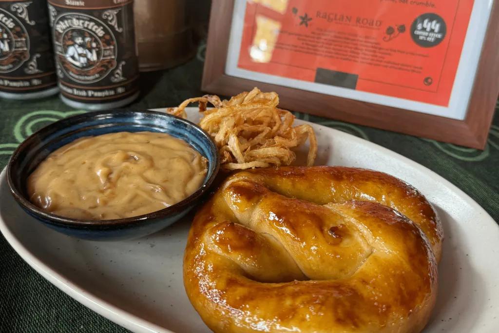 Potato Pretzel with a yellow mustard looking sauce and crispy food on the side. There are Oktoberfest cans in the background with beer in them.