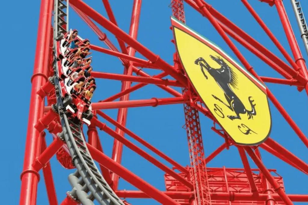 Big tall rollercoaster which goes straight up and falls down steep drop. There is a huge Ferrari badge, a yellow shield with a black silhouette of a horse and the initials S and F. The rollercoaster car is shaped like a red Ferrari. 