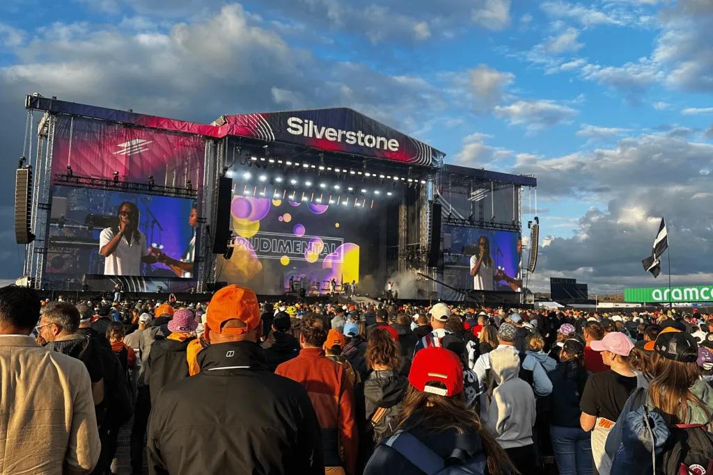 There is a large stage with multi-coloured lighting on it. There is a large crowd watching the stage wearing different F1 teams merch. The stage says Silverstone at the top of it.