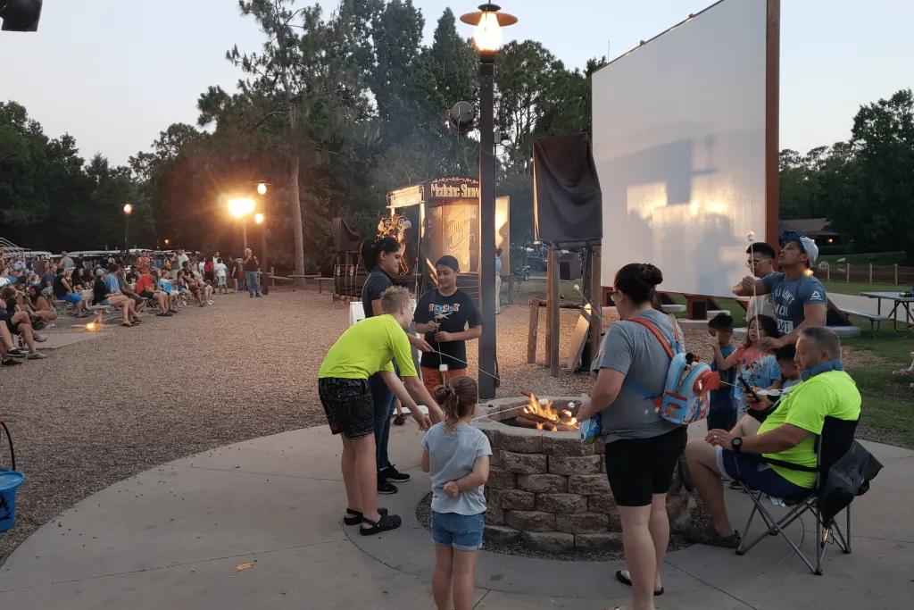 Guests are gathered around a campfire roasting marshmallows on sticks. There are many tables full of guests and the sun is setting over the camp site. There is a little stage for Chip 'n' Dale to perform on and a board for movies to be projected onto.