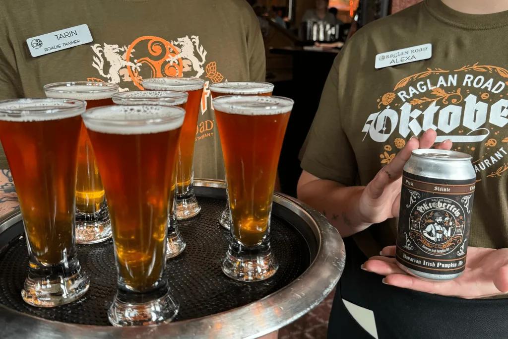Two workers holding drinks and wearing dark green t-shirts which say Oktoberfest and have nametags on. One is holding a black tray with large shots on and the other is holding a silver can which has Oktoberfest theming on it.