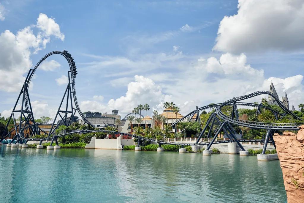 A pristine blue lake with a tall rollercoaster towering in the background. It is grey and features a huge drop and many twists and turns.