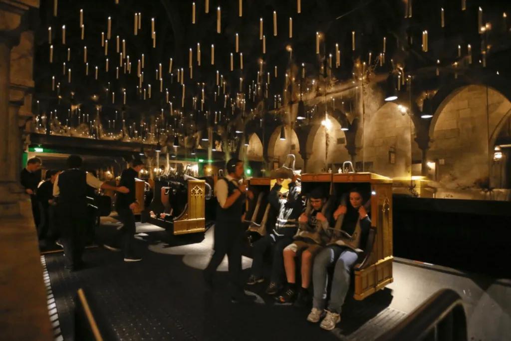 A dark room with floating candles over head. There are attendants holding torches as they strap guests into the ride vehicle safely. There are four chairs in a row.