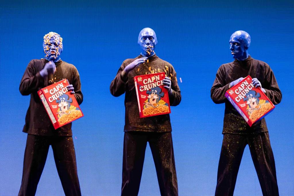 Three men wearing all black outfits stood on stage painted completely blue. Each are holding boxes of Captain Crunch cereal. From the left to the right they progressively have more and more cereal stuck to them, with the man on the left having pieces all over his head. 