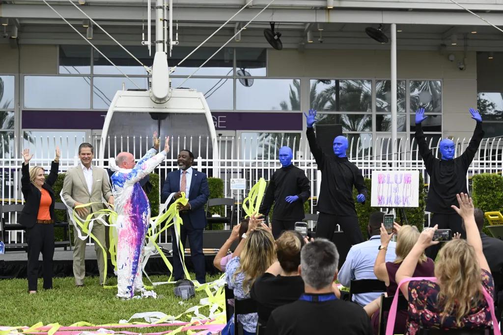 Three men dressed in all black and painted completely bright blue stood with their arms up looking at a man in an all white suit with paint splattered all over him. Behind there are Ferris wheel carriages at the station and there are many people sat in rows of seats cheering along.