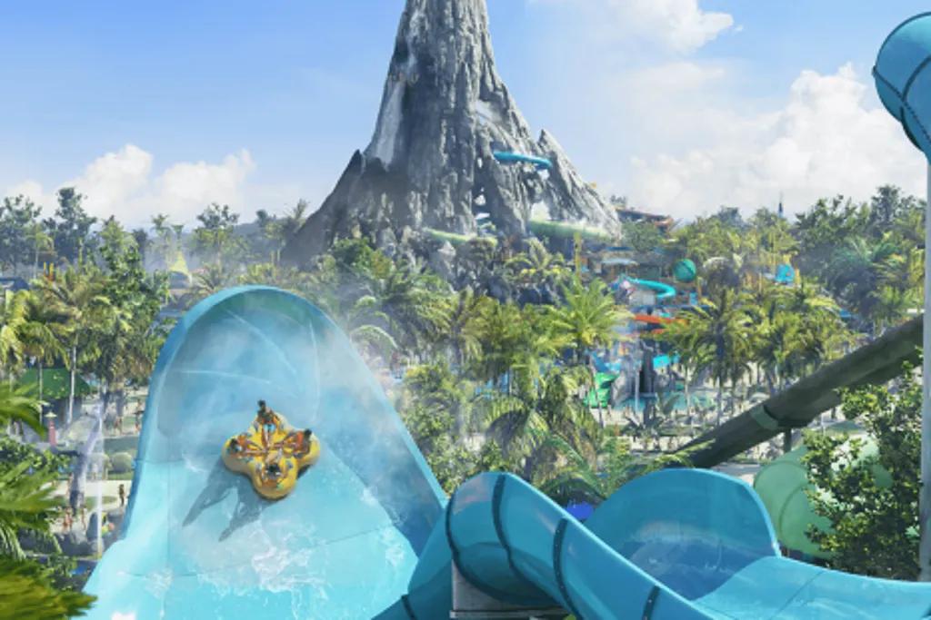 Four people facing each other on a large yellow rubber ring flying up a light blue water slide wall which is huge. In the background, there is a volcano statue with slides coming off of it and water.