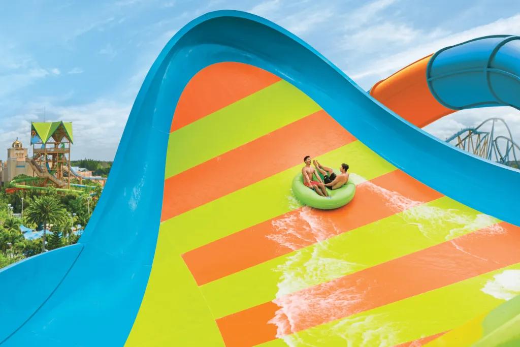 Two men sat on two seater lime green rubber ring on a blue slide with lime green and orange stripes. They are heading up a large curled up wall.