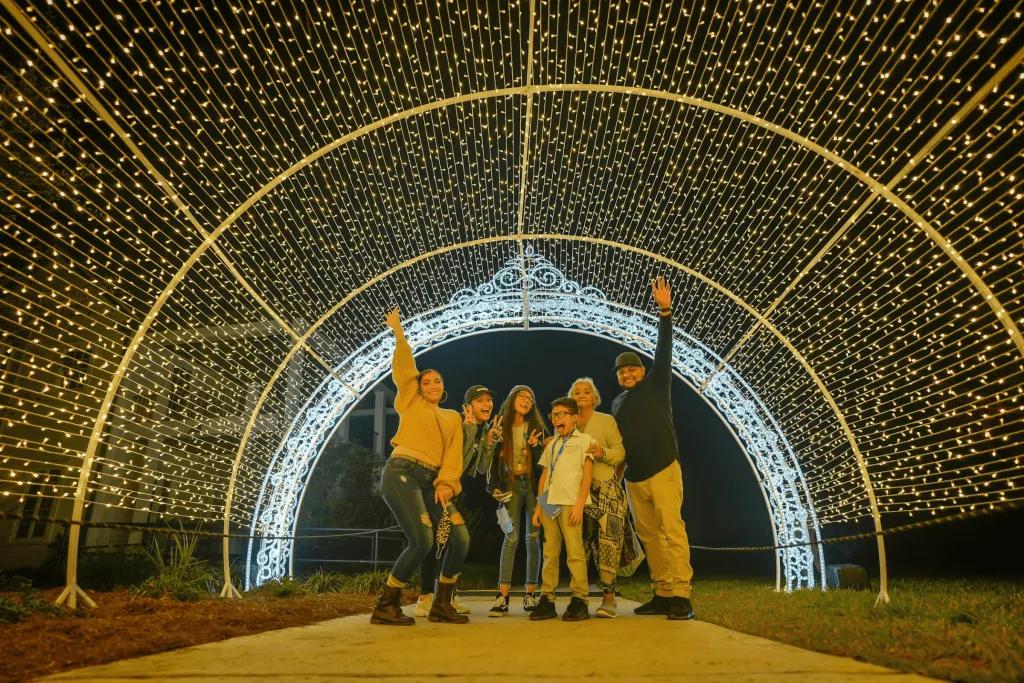 Family of 6 stood under a tunnel of lights at night. They have their arms lifted up towards the magical yellow and white lights.