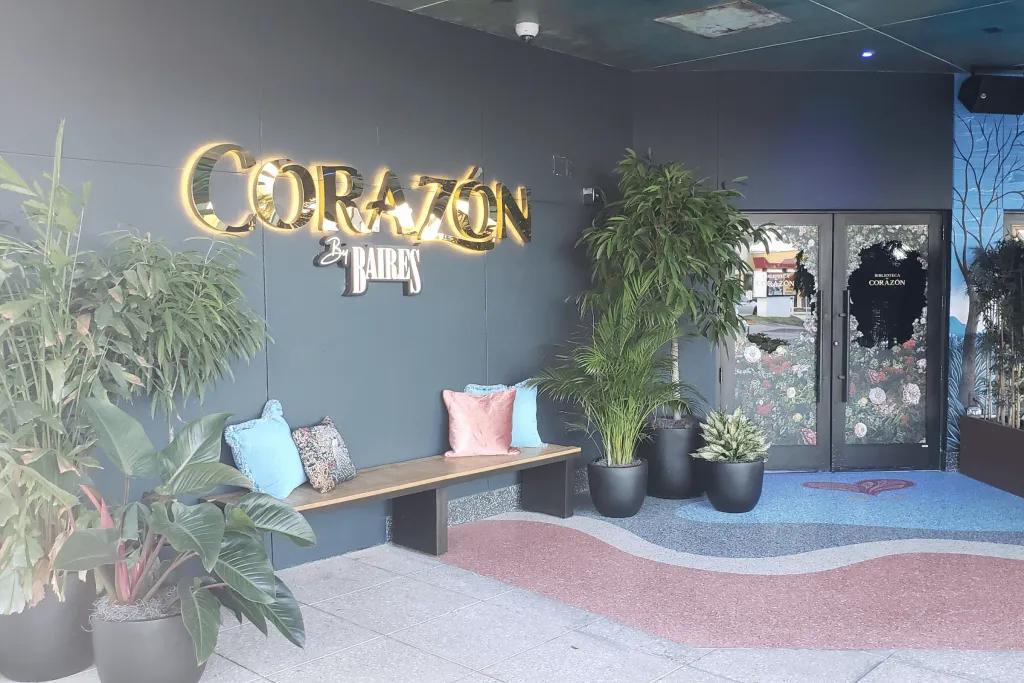The entrance to the restaurant with a golden sign saying corazon lit up on the dark grey wall underneath there is a wooden bench with pillows. To the right there are window door.