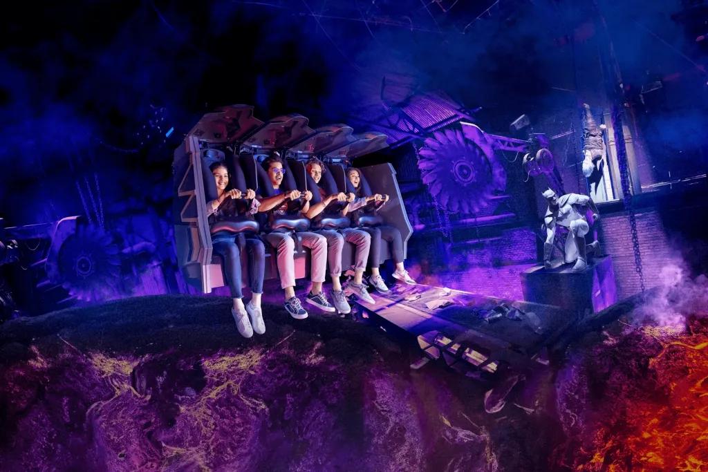 Group of four sat on rollercoaster car held in by lap bars with their legs hanging. In the background you can see Batman sat in a dark and dingy area.