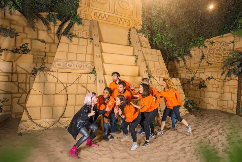 Woman wearing wacky outfit and pink wig is crouching with a group of guests who are wearing matching orange jumpers. They are stood on sand are in front of two large temple.