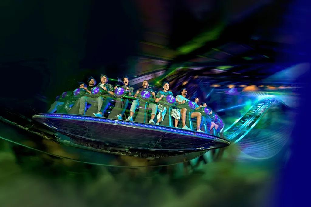 People sat on circular vehicle facing outwards and spinning, strapped onto vehicle with back restraint. The colour scheme is blue and green.