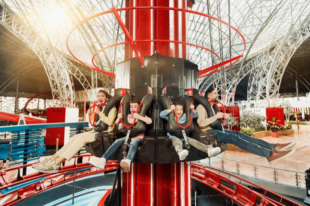 A circular drop tower inside of a large industrial white building. There are small kids and adults on the drop tower smiling and screaming.