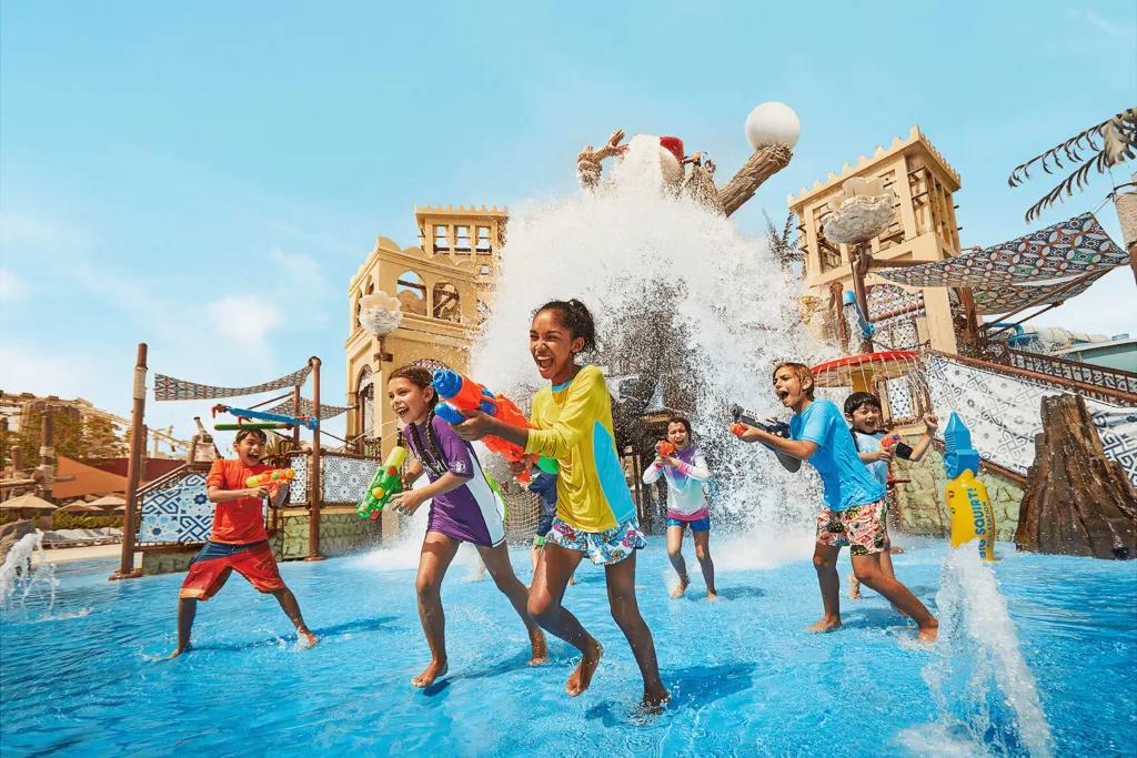 There are many little kids running away from play area large bucket pouring huge wave of water down onto them. They are all holding water guns and laughing pointing them at one another. The play area has a large climbing frame in a shallow pool.