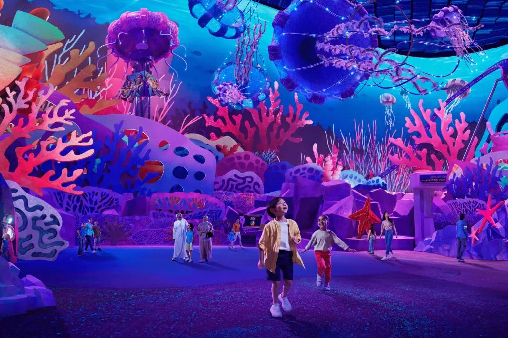 Young boy walking through a large room with luminous decorations in it. There's large coral reef decorations and flying overhead are hanging sea weed decorations.