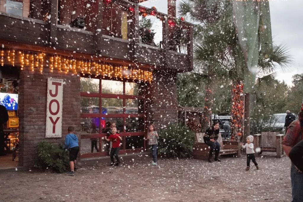 Stone building with Christmas decorations including orange lights and a sign saying JOY on it. There is fake snow floating down and young kids playing in it.