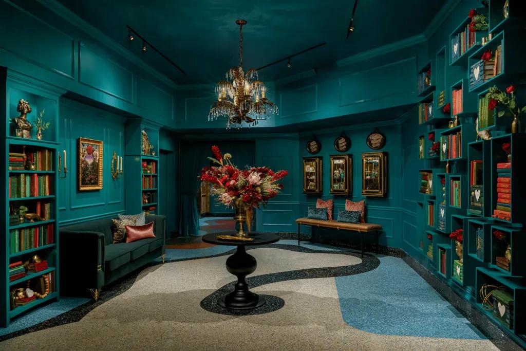 Turquoise large room with many shelves full of books on the wall, an obscure white, blue and black swirling pattern. In the centre there are flowers in a large vase.