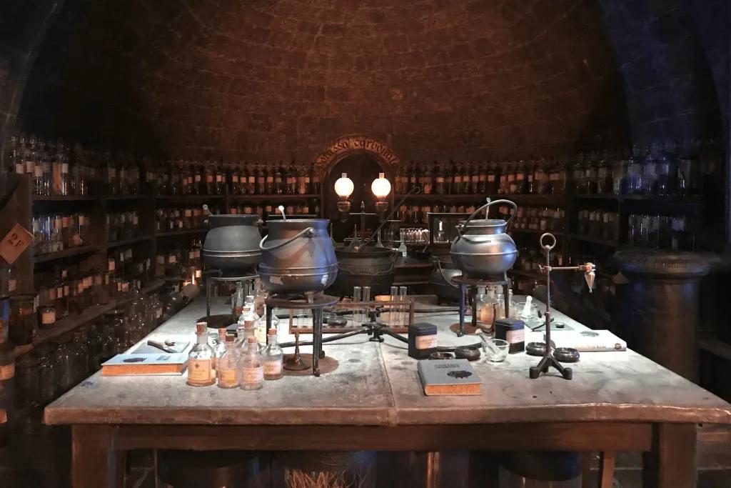 A dark lit room with a dimly lit main table with multiple black cauldrons and substances. In the background there are dark shelves holding test tubes with different potions and substances.