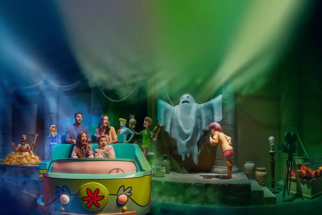 A family of four are sat in a ride vehicle with two rows of seats painted like the car from Scooby Doo. In the background there is the gang from Scooby Doo and Velma is whispering in the ear of a ghost with its arms held in the air.