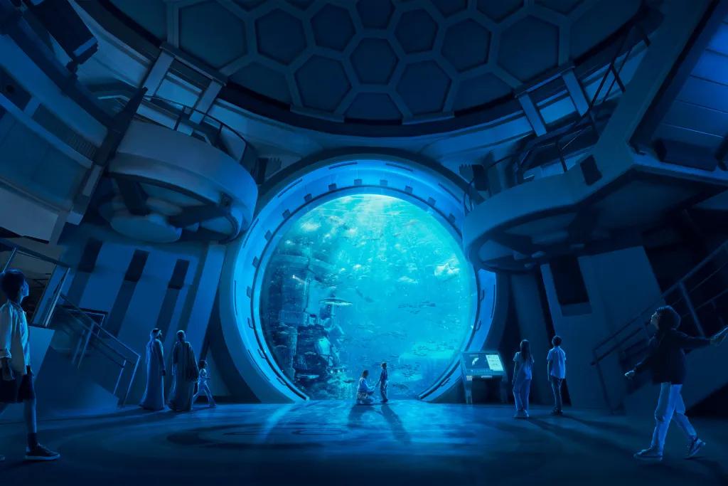 Mum kneeling and holding hand of child stood in front of a huge circular window. On the other side there are fish and marine animals swimming around. The room is dark and there are a lot of guests walking around.