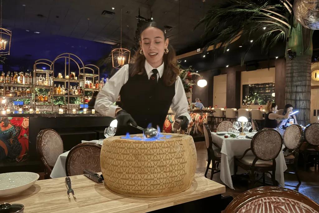 Waitress dressed in black and white suit and waistcoat dishing food in a giant wheel cheese and holding a large spoon which is lit on fire. In the background, you can see the classy decor of the restaurant with the bar and palm trees lit with sparkling lights.