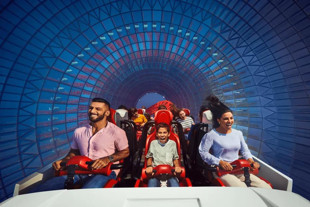 A row of three guests sat on a white roller coaster car with red lap bars holding them in. There is a tunnel surrounding coloured blue and they are all screaming with excitement.