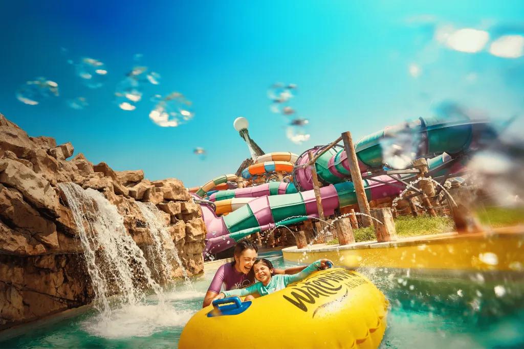 In the background there are many large waterslides wrapping around one another in different colours. There is a small lazy river flowing with a waterfall pouring into it off of rocks. There is a mother holding her little girl who is sat on a yellow rubber ring. There are fountains also splashing them.