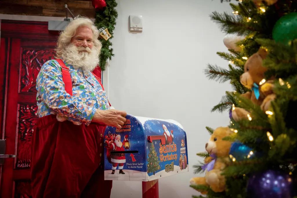 Santa, a man with blue patterned shirt on and red dungarees on over the top made of velvet. He has white hair and a white beard. He is holding a gift and putting it under the Christmas tree, decorated in gold and blue decorations.