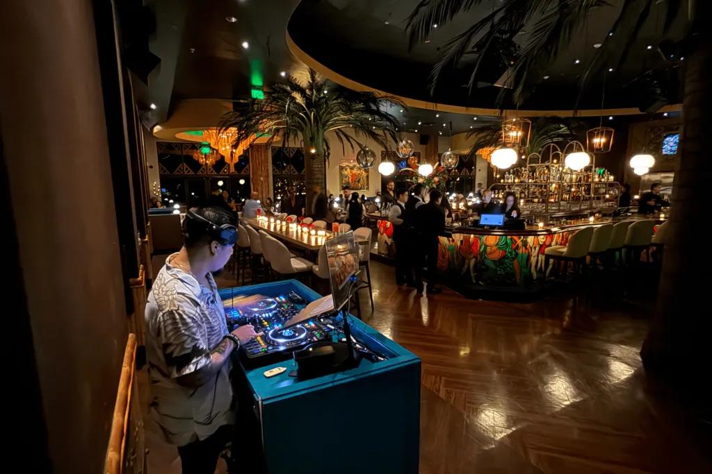 The dimly lit restaurant with a man stood behind a turquoise dj deck wearing headphones. In front there is a small dancefloor space and the tables are in the background.