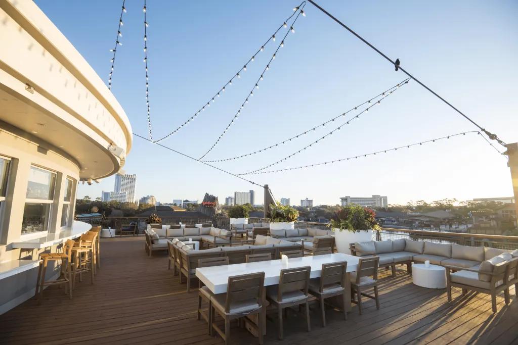 A wooden terrace outside of a restaurant with lights hanging over. There are many tables with chairs on this terrace.