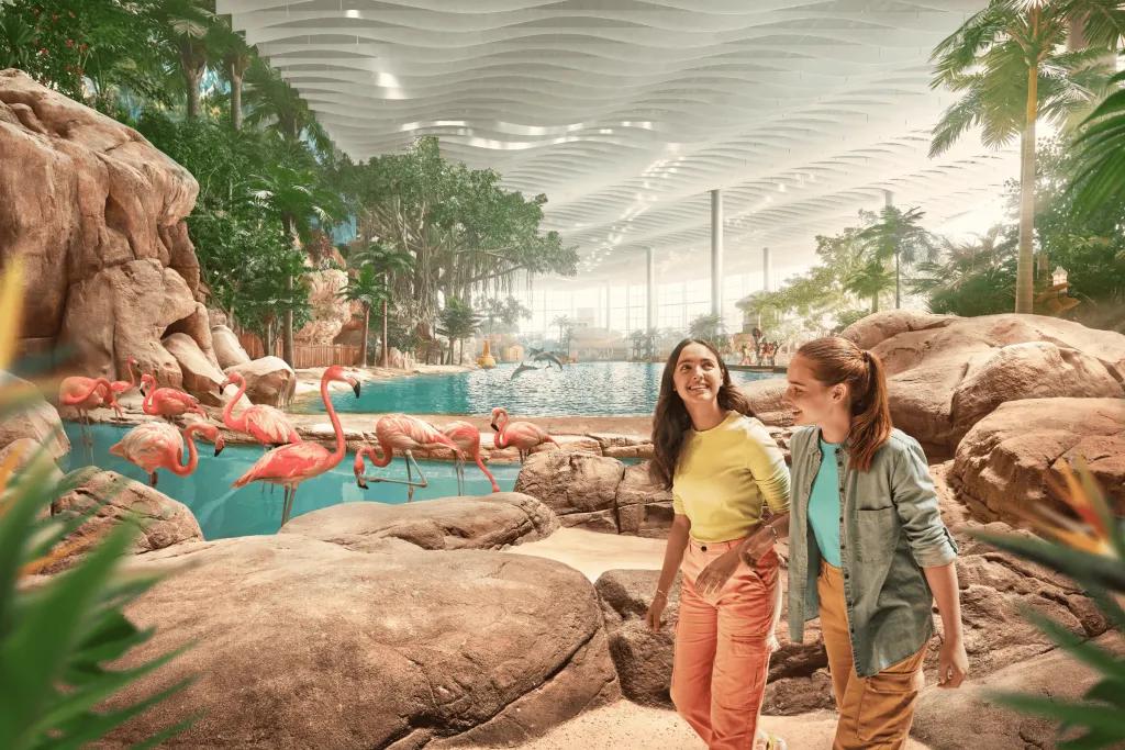 Two young women walking along a rock pathway with pools inside a building. There are flamingos stood in the pool. 