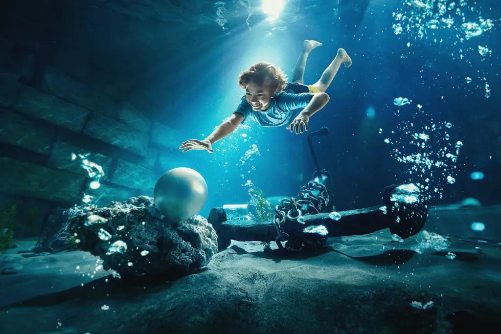There is a young woman diving down into a deep pool reaching out to a large white pearl sat at the bottom of the pool. There is also an anchor sat at the bottom of the pool and bubbles surrounding the woman.