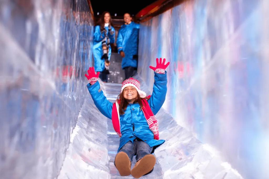 Slide made of ice with a young girl wearing a long blue jacket, red santa hat and red gloves. She is smiling with her arms lifted in the air.