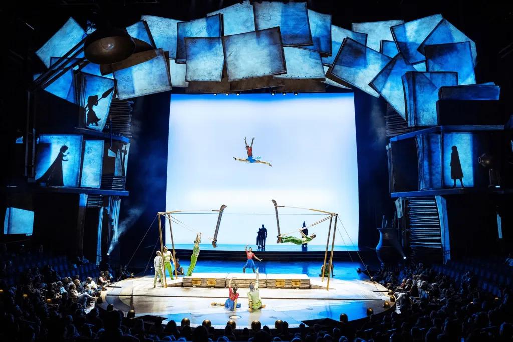 A large stage lit in blue lights and incredible theming with pages surrounding it. There are gymnasts on the stage with one leaping in the air in the splits.
