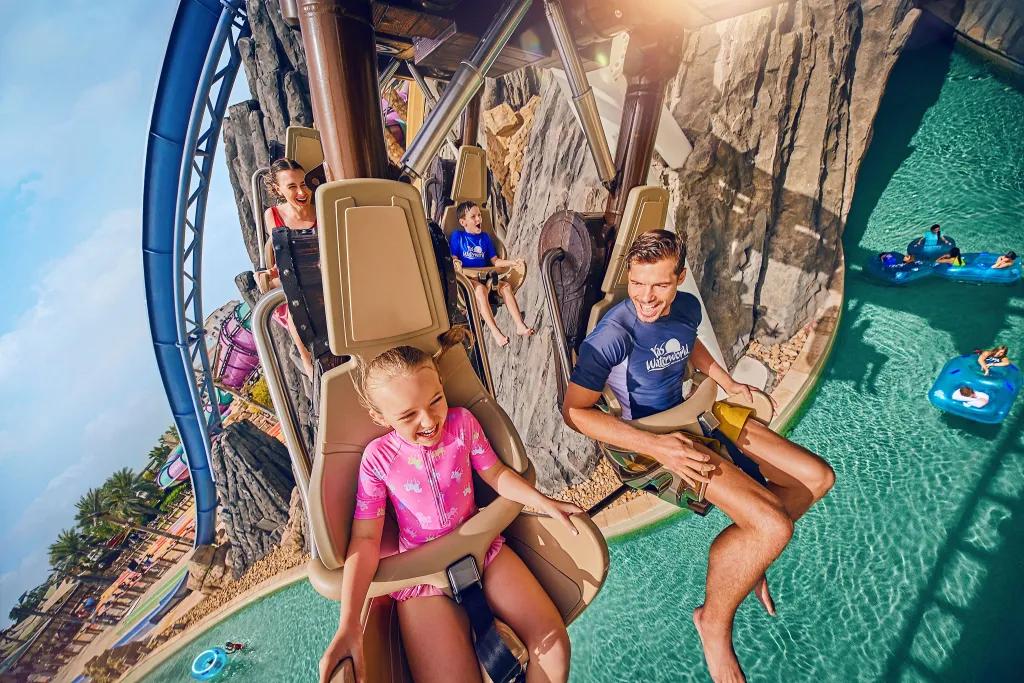 A child and her dad sat on a hanging coaster in their swimwear smiling and laughing as they glide above the large swimming pool. Below there are people lounging on rubber rings in the lazy river.