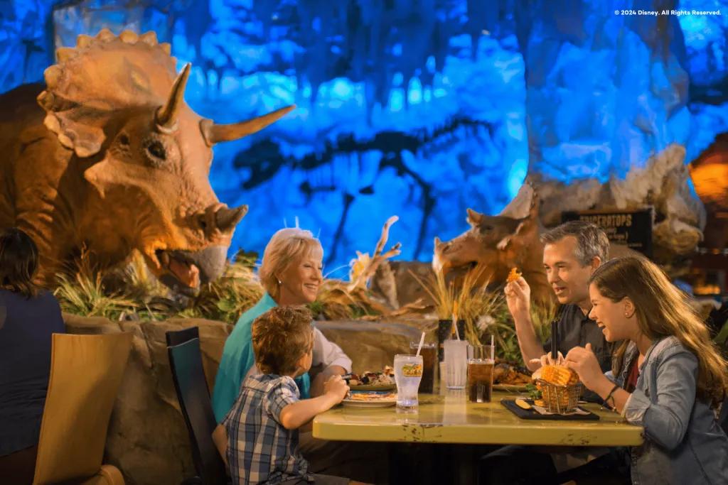 Grandparents and two children laughing eating burgers and fries with dinosaur statues and skeletons behind them on display.