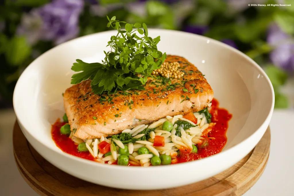 Beautiful dish of salmon presented with leaves on top and ride and peas below and a red tomato sauce below on a bowl. Behind there is a bush.