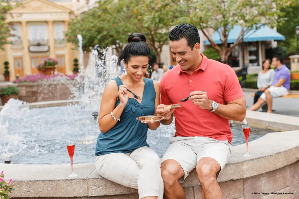 Couple dining sat on the stone edge of the fountain in the French Pavilion in the world showcase. They have small plates of food and glasses of sparkling red drinks. They are both smiling.