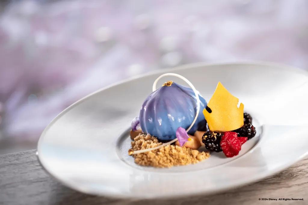 Blue and purple droplet shaped dessert with gold on top. There is also biscuit crumbs at the bottom, fruit beside and a golden crown on top.