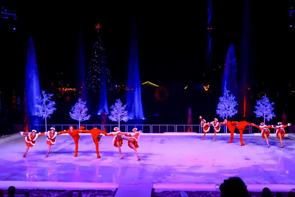 People skating on large ice rink in Christmas outfits all in a line with leg lifted up. Behind the rink there are small Christmas trees lit in blue lighting.