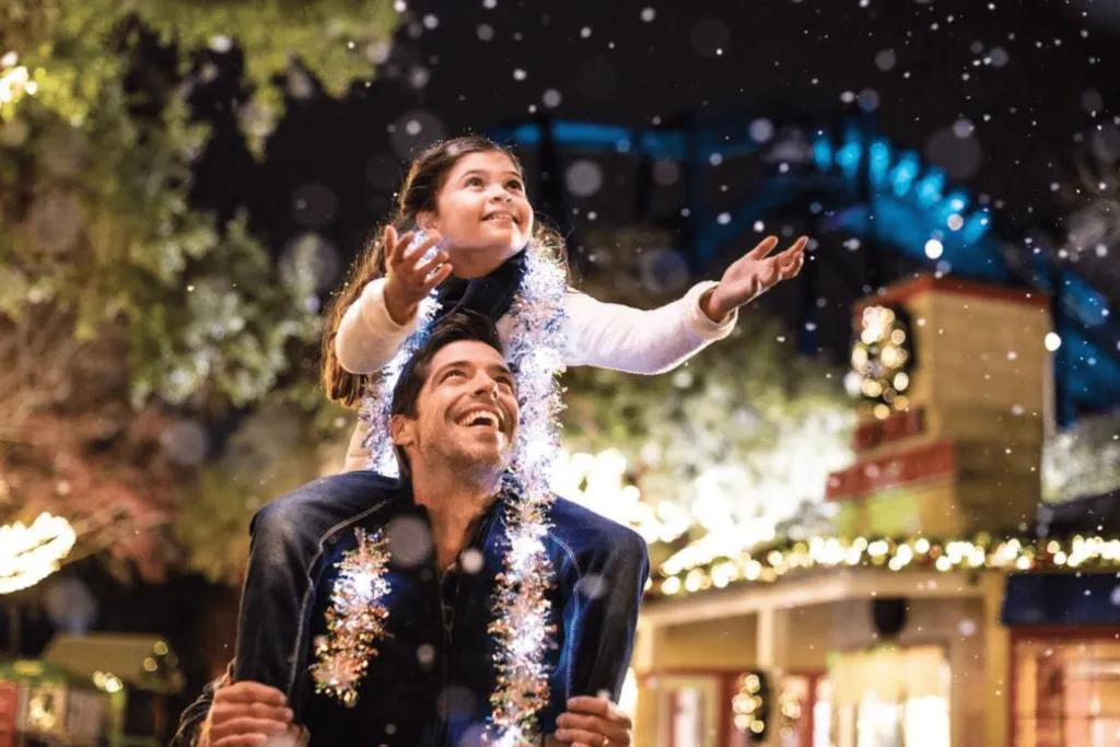 Dad looking up at his daughter who is sat on his shoulders and wearing tinsel around her neck. The little girl is reaching out to touch the fake snow. Behind there are trees and buildings decorated with lights and reefs.